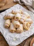 A pile of Grandma's Christmas nut horns, rolled crescent cookies with walnut filling, dusted with powdered sugar on parchment paper