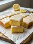 Moist Lemon Sheet Cake 6 Squares of moist lemon sheet cake topped with lemon glaze and lemon zest, stacked on parchment paper with fresh lemons