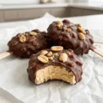 Chocolate-coated peanut butter frozen yogurt bars on sticks topped with chopped peanuts, one with a bite taken out