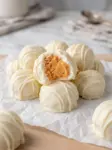 A stack of peanut butter snowballs coated in white chocolate with drizzle, one bitten to show the creamy peanut butter center