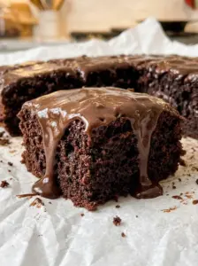 A square slice of triple chocolate cake with hot fudge sauce dripping down the side of the moist chocolate cake